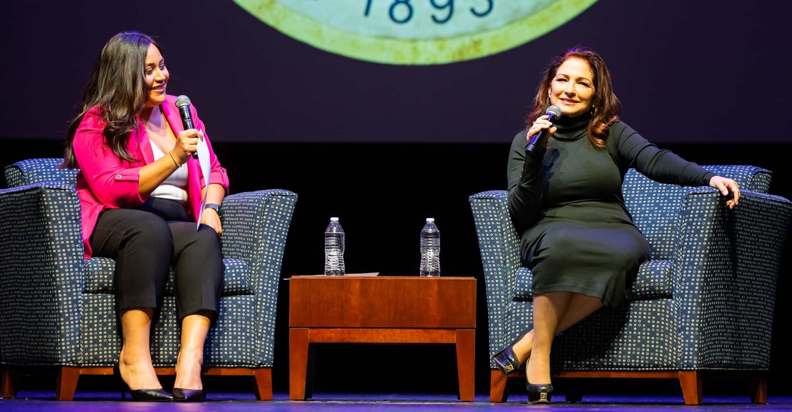 Briceyda Landaverde and Gloria Estefan are seated in chairs on the Lyman Center stage and Gloria is speaking to the audience
