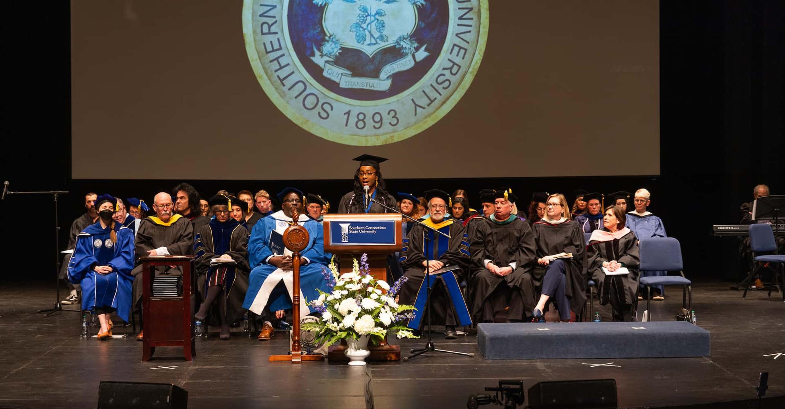 faculty and administrators in academic regalia sit on the Lyman Center stage as a student wearing a graduation cap and gown speaks at the podium