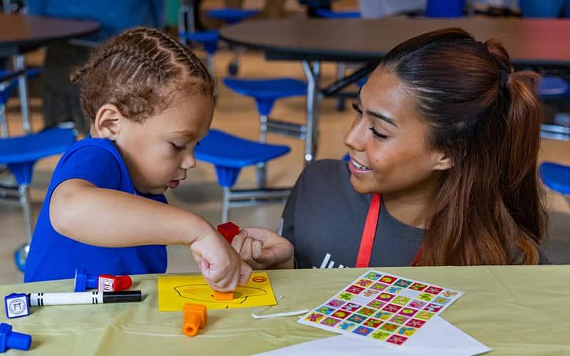 a preschool teacher and a young child work together on a small art project