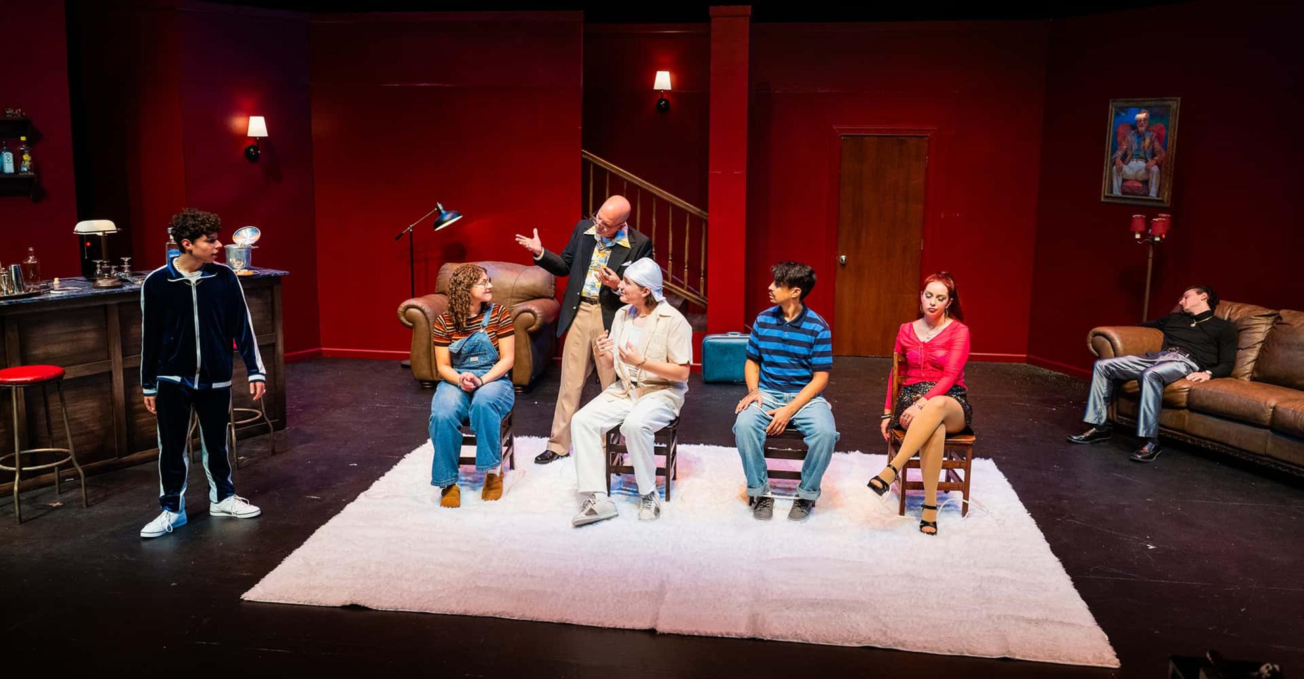 four people sit on chairs in a room, with two other people standing and one other person on a couch