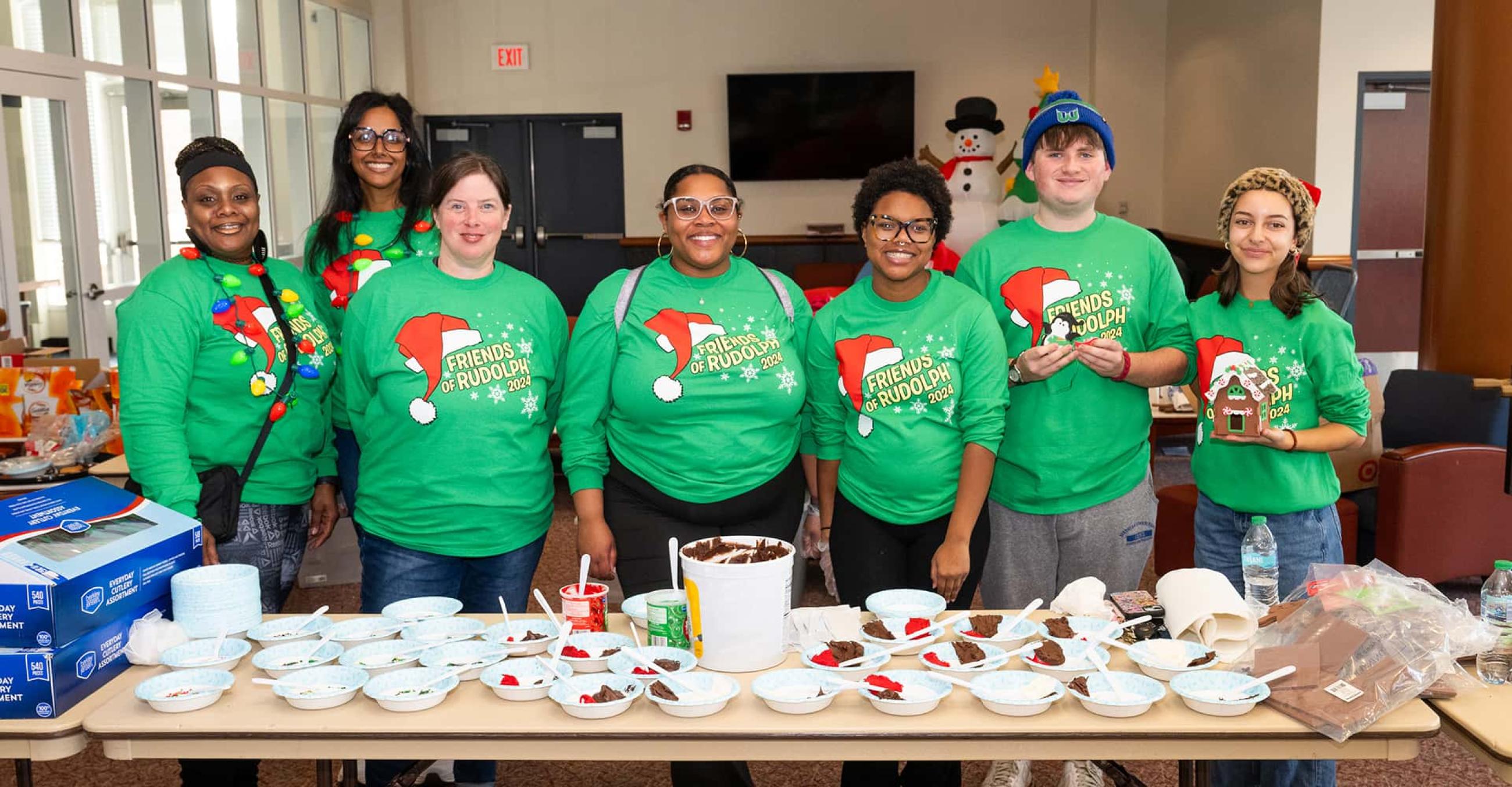 a group of 7 volunteers wearing green "Friends of Rudolph" shirts stand behind a craft table