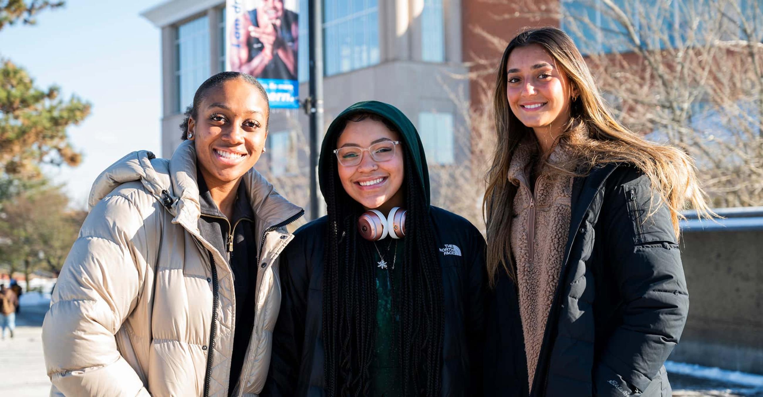 three smiling students in winter jackets stand in front of the student center